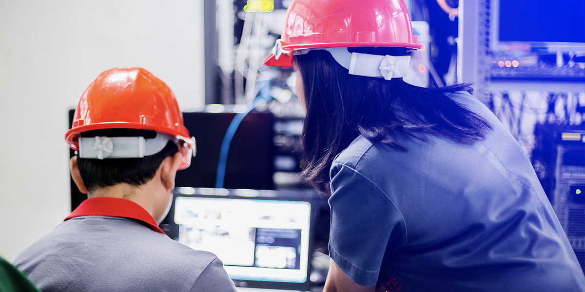 two employees in hardhats conversing in front of a computer